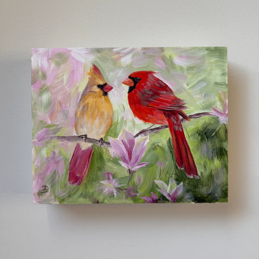 An 8x10 painting of two cardinals perched on branches among pink flowers, mounted on a light gray wall.