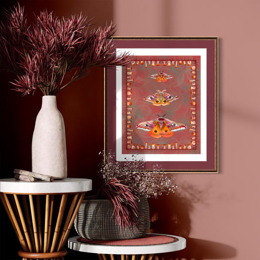 A framed art print featuring three colorful moths on a textured red background, styled in a warm, rustic interior setting with dried plants and decorative tables.
