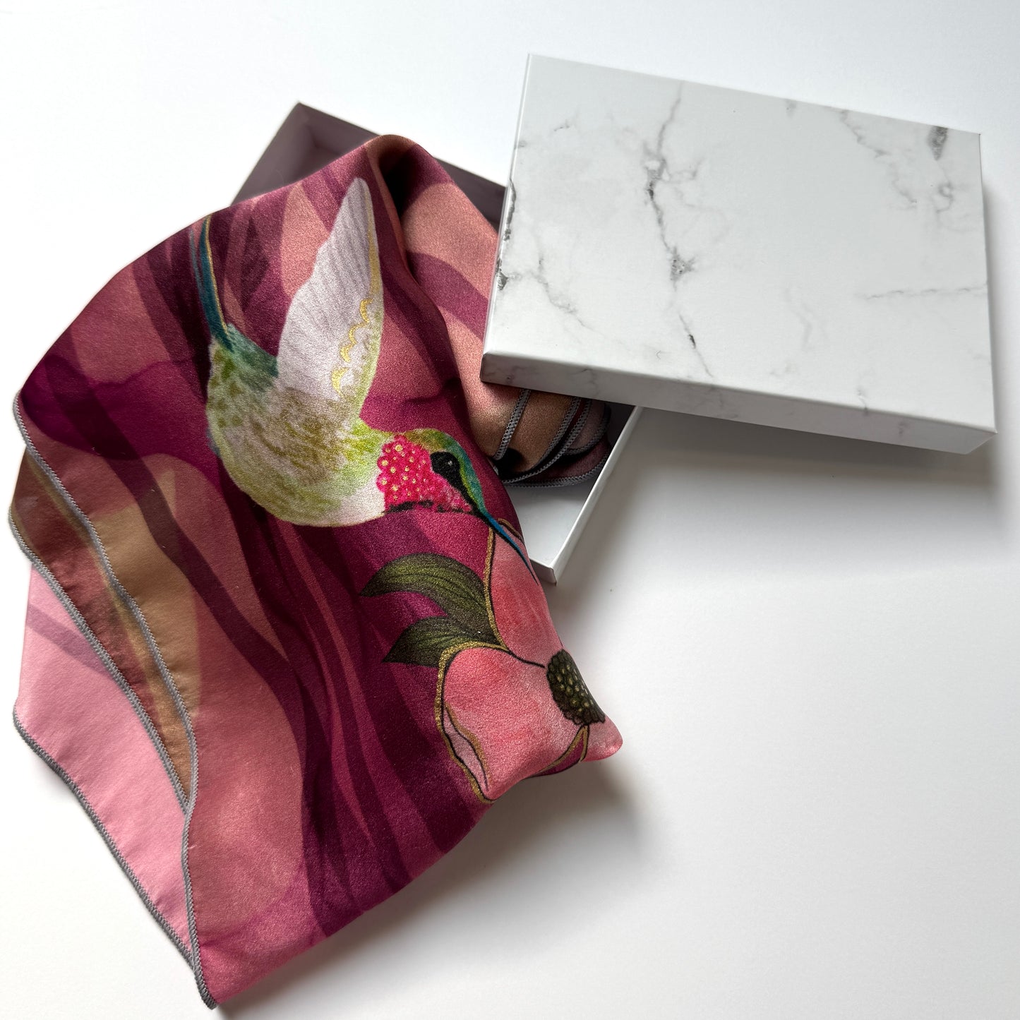 Decorative scarf with floral and hummingbird design on a white surface next to a marble-patterned box. For organizations seeking luxury corporate gifts and unique client appreciation gifts.