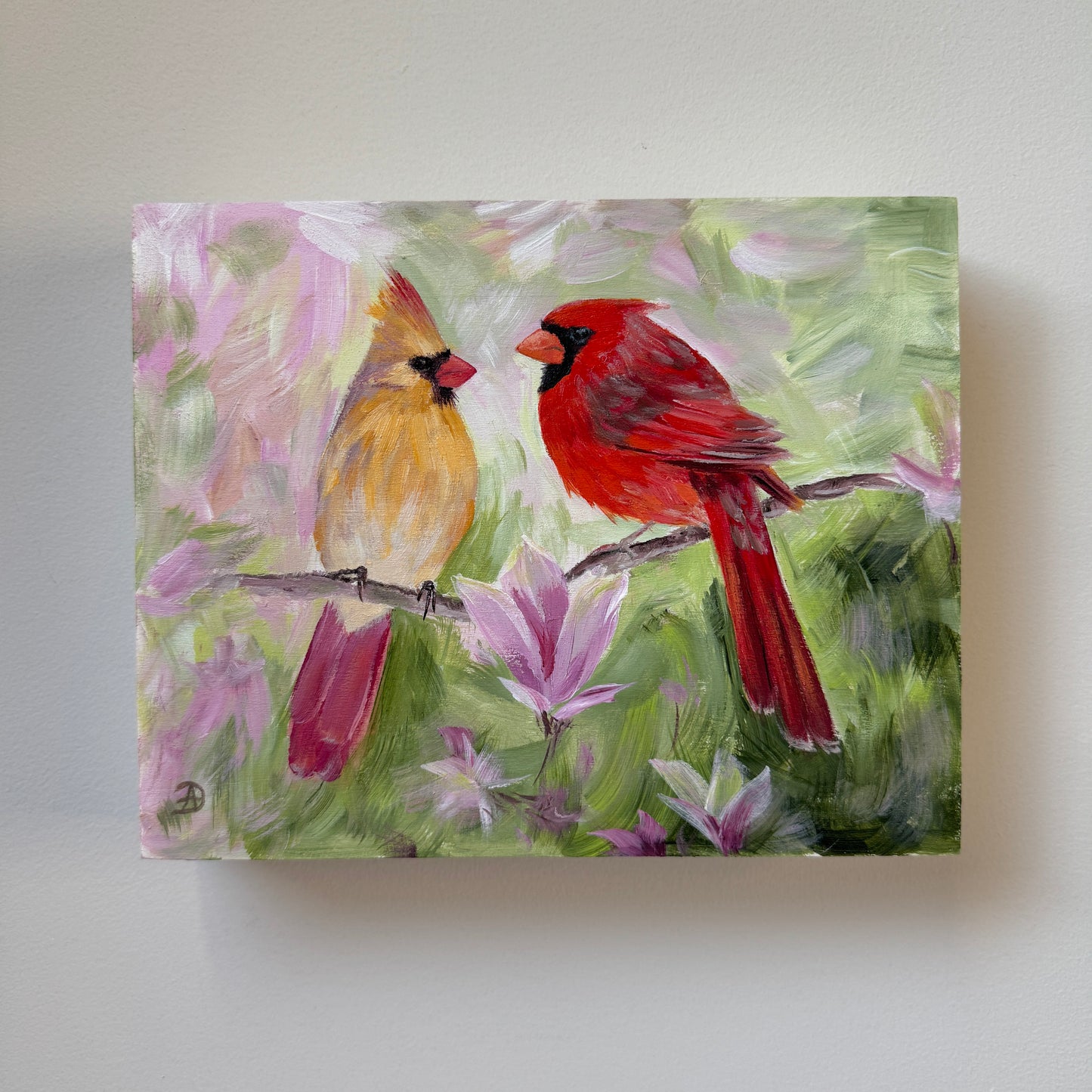 An 8x10 painting of two cardinals perched on branches among pink flowers, mounted on a light gray wall.