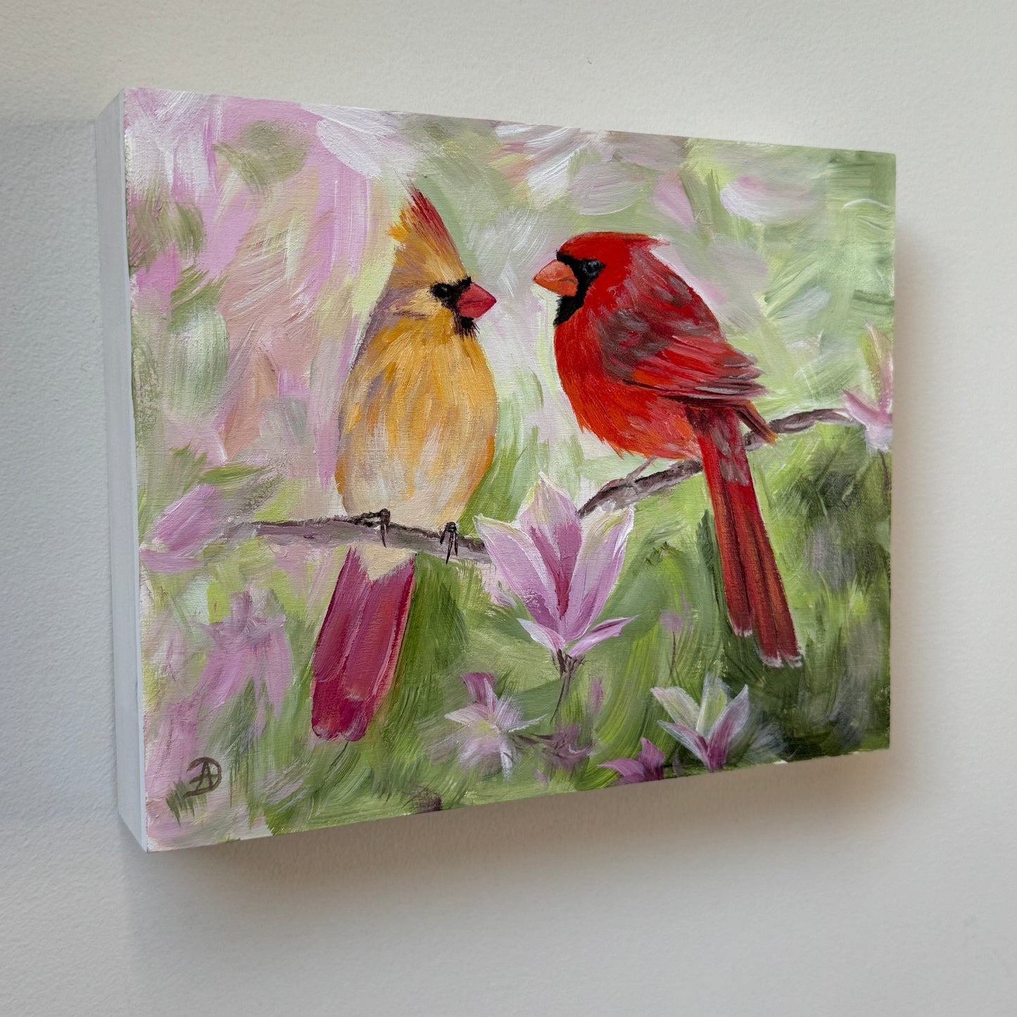 An 8x10" painting of two cardinals perched on a branch, with soft pink magnolia flowers and green foliage in the background, mounted on a light gray wall. 