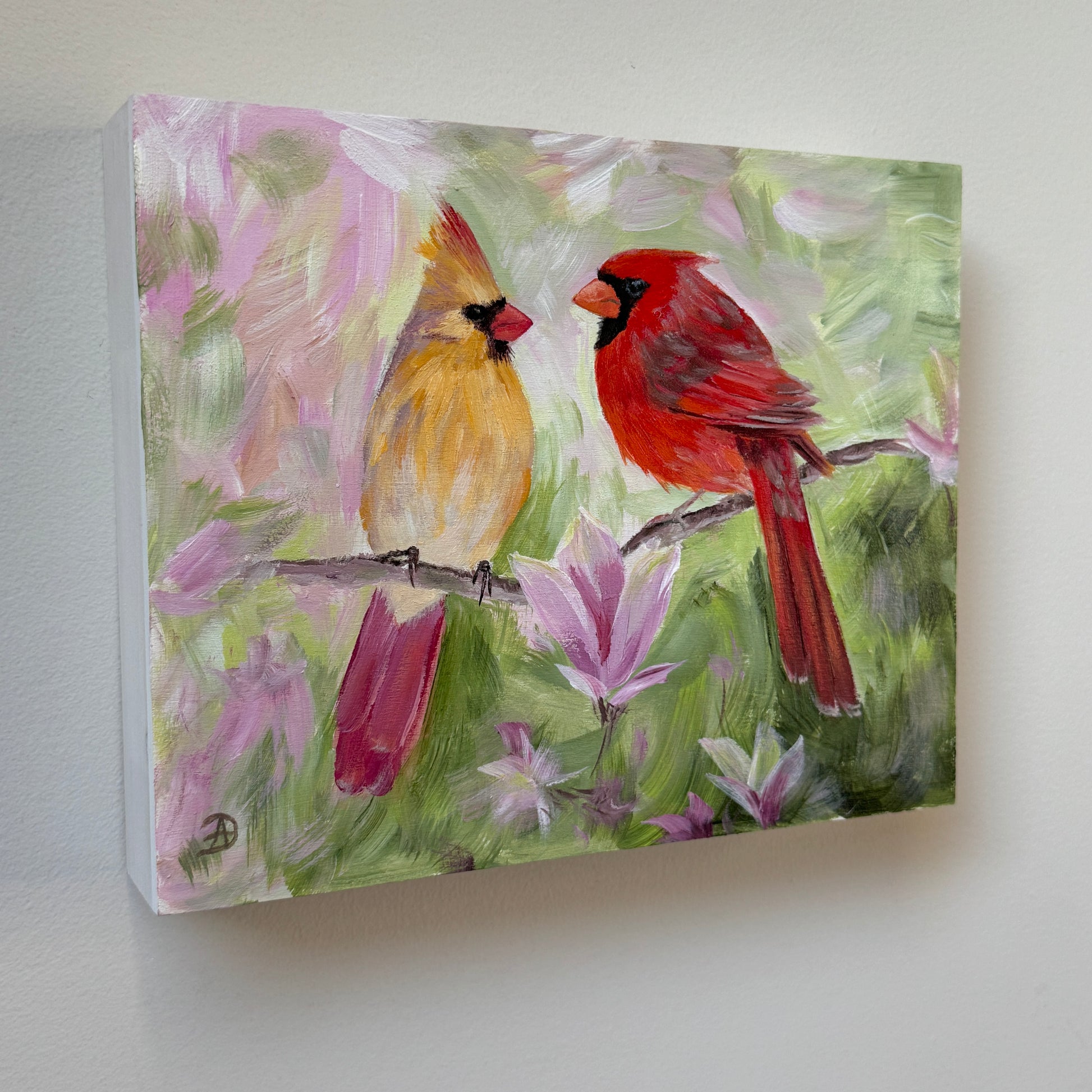 An 8x10" painting of two cardinals perched on a branch, with soft pink magnolia flowers and green foliage in the background, mounted on a light gray wall. 