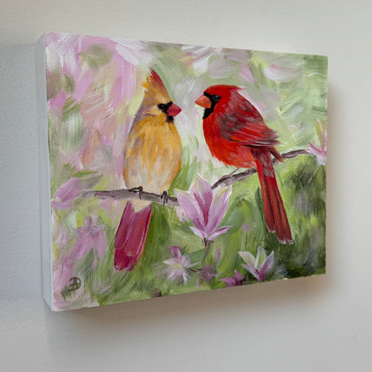 An 8x10" painting of two cardinals perched on a branch, with soft pink magnolia flowers and green foliage in the background, mounted on a light gray wall. 