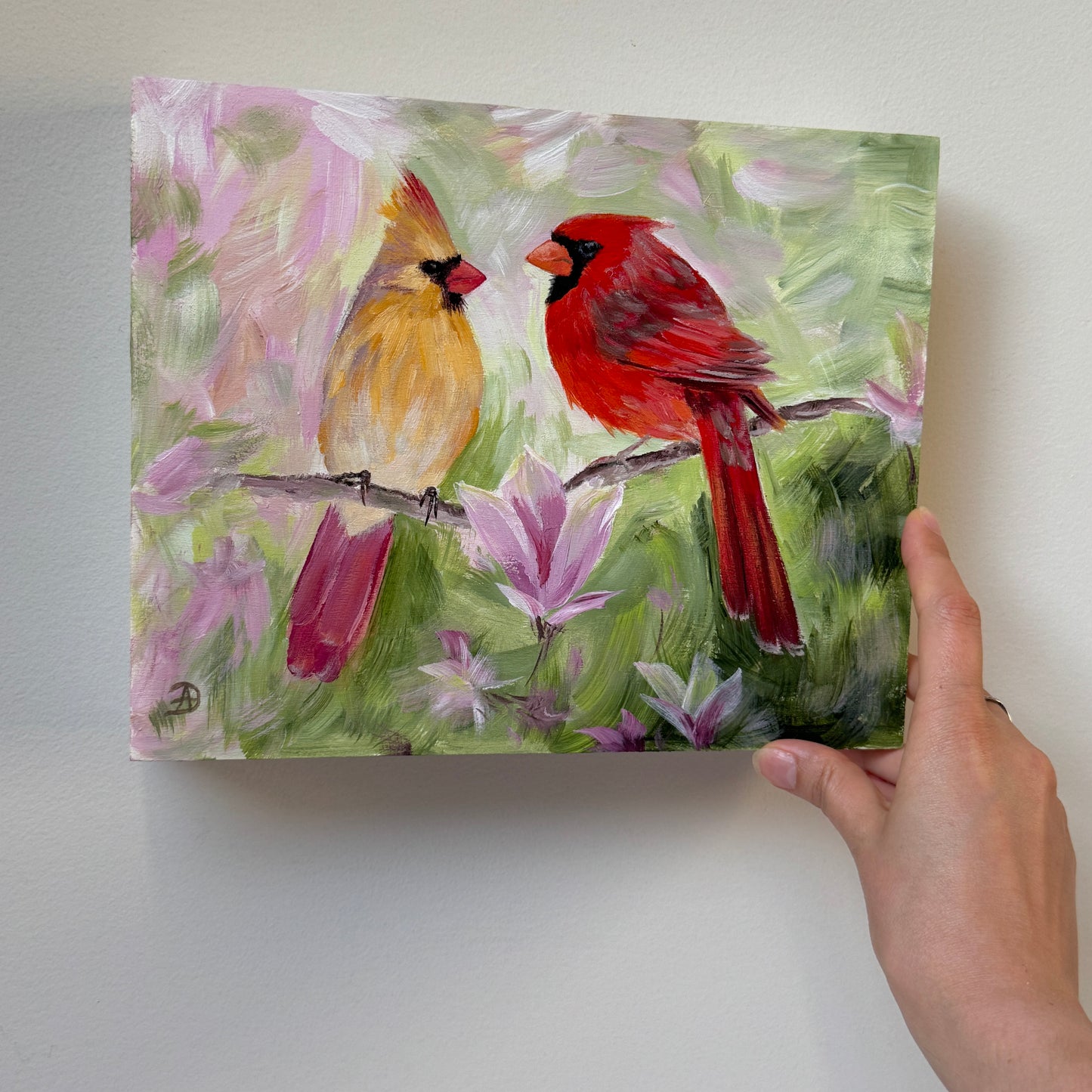A hand holding an 8x10" painting of two cardinals perched on a branch among pink magnolia flowers, shown against a plain light gray wall.