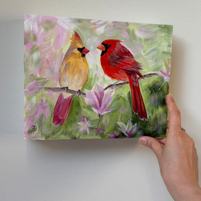 A hand holding an 8x10" painting of two cardinals perched on a branch among pink magnolia flowers, shown against a plain light gray wall.