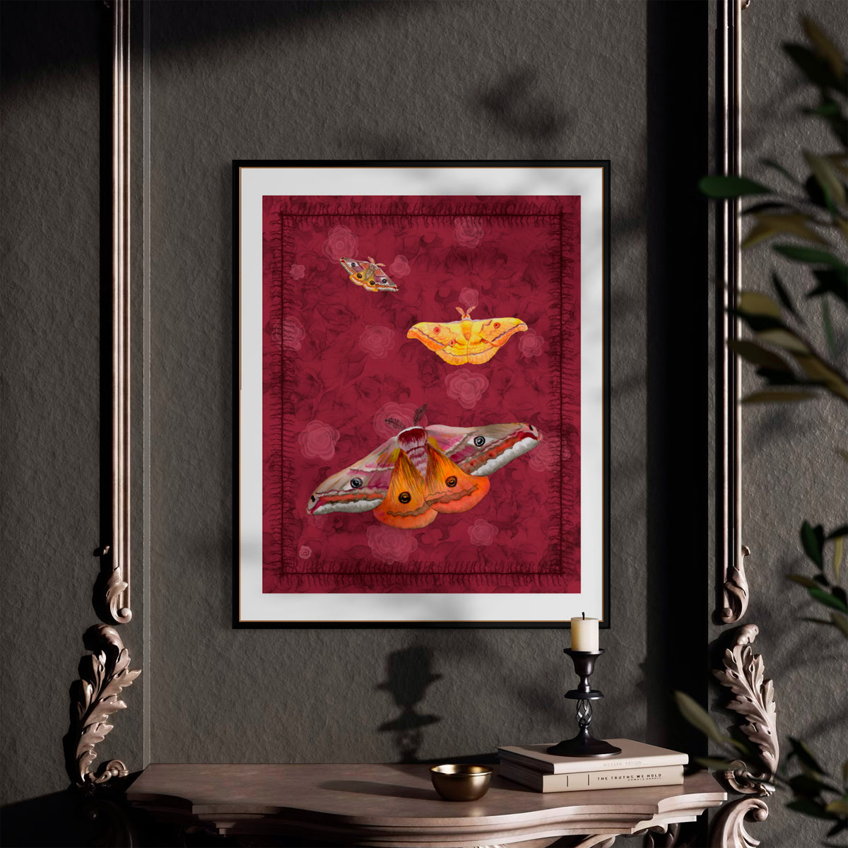 A framed art print featuring three stylized moths on a textured deep red background, displayed on a dark gray wall above a console table with a candle and books.