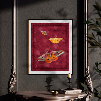 A framed art print featuring three stylized moths on a textured deep red background, displayed on a dark gray wall above a console table with a candle and books.