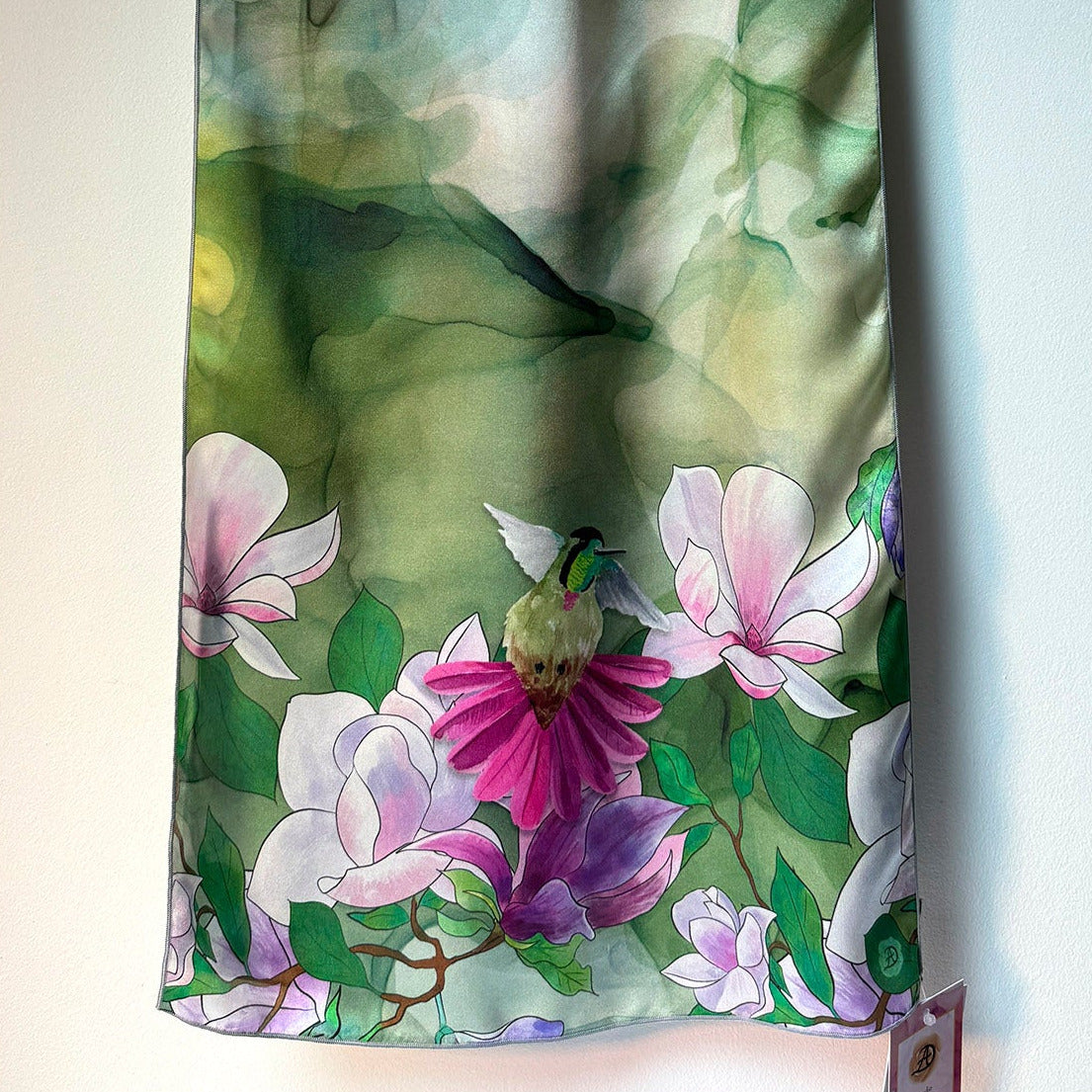 Floral scarf with a hummingbird and pink magnolia on a green background, hanging against a white wall.